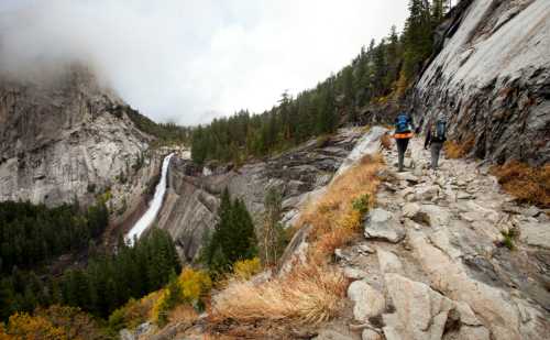 Two hikers walk along a rocky trail beside a waterfall, surrounded by lush trees and misty mountains.