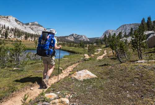 A hiker with a backpack walks along a trail beside a serene lake, surrounded by mountains and trees under a clear blue sky.