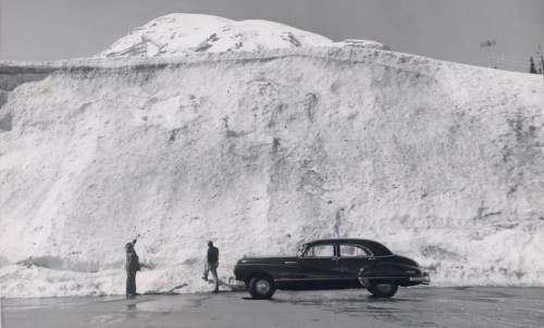 Two men stand near a vintage car beside a massive snowbank, with a mountain in the background.