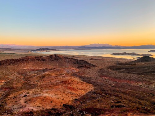 A scenic view of a desert landscape at sunset, with mountains and a calm lake in the background.