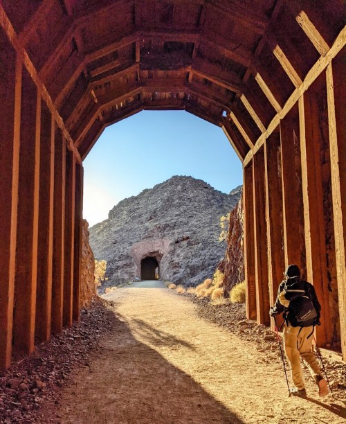 A person walks through a rustic tunnel, leading to a mountain view and another tunnel in the distance.