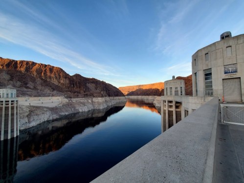 A serene view of a dam with mountains in the background, reflecting in calm water under a clear blue sky.