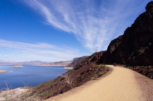 A scenic path along a lake, with two hikers walking beside rocky cliffs under a blue sky with wispy clouds.