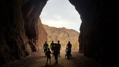 Silhouetted figures walking through a rocky tunnel, with a scenic landscape visible in the background.