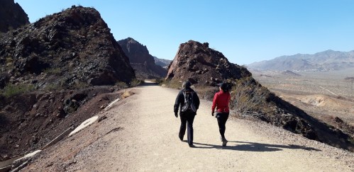 Two hikers walking on a dirt path between rocky hills in a desert landscape under a clear blue sky.