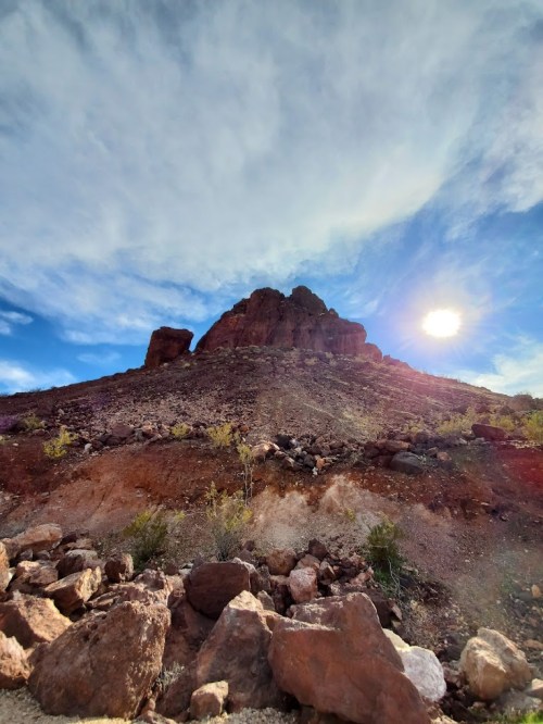 A rocky hillside under a blue sky with scattered clouds and the sun shining in the background.