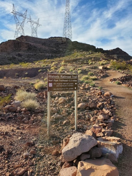 Sign for the Historic Railroad Trail, with a rocky path leading up a hill and power lines in the background.