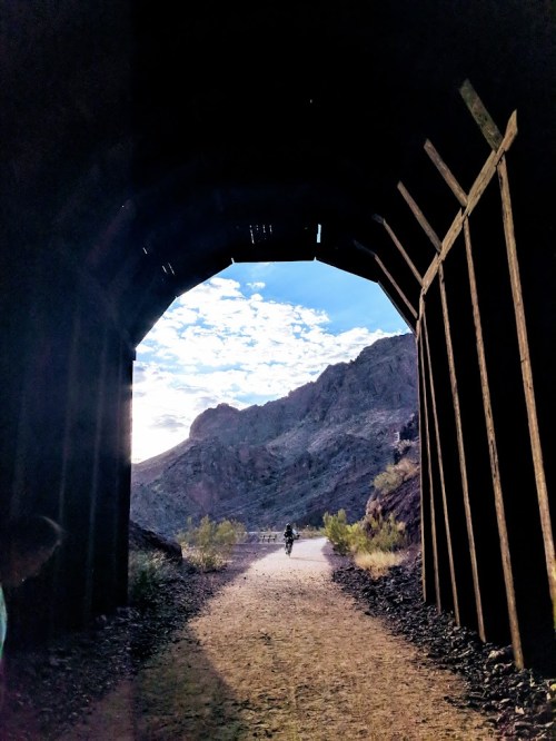 A cyclist rides through a dark tunnel, with mountains and a bright sky visible in the background.
