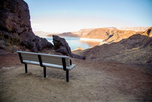 A solitary bench overlooks a serene lake surrounded by rocky hills and mountains under a clear sky.