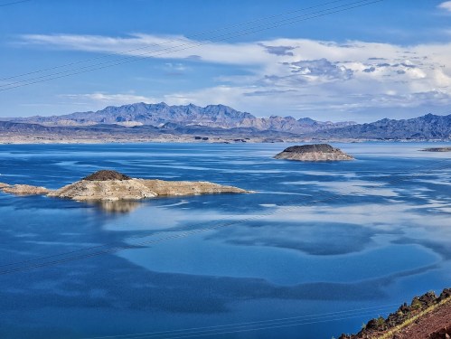 A serene lake with two small islands, surrounded by mountains and a clear blue sky. Reflections shimmer on the water.