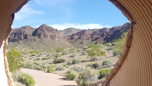 View of rugged mountains and desert landscape through a curved, metal structure. Clear blue sky above.
