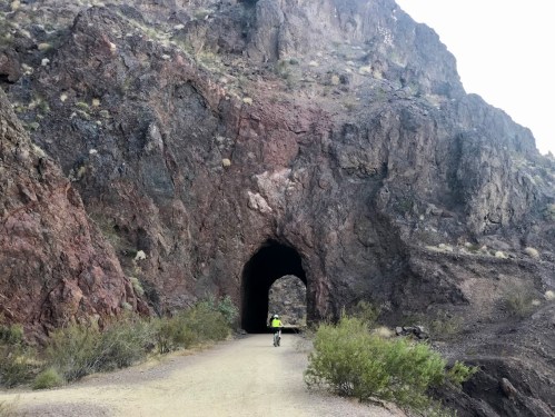 A cyclist approaches a dark tunnel carved into a rocky mountainside, surrounded by desert vegetation.