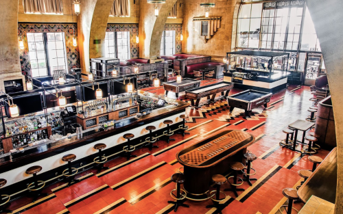 A spacious bar interior with wooden furniture, pool tables, and a vibrant floor design, featuring warm lighting.