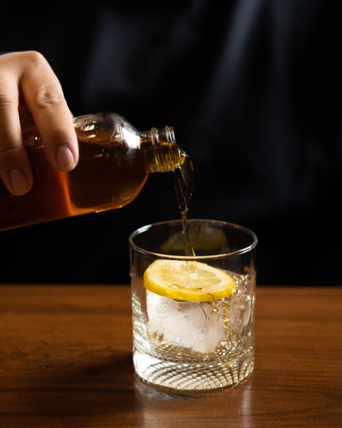 A hand pours a dark liquid into a glass with ice and a lemon slice on a wooden table.