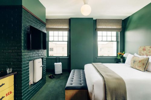Cozy hotel room with a green accent wall, a bed, a bench, a TV, and large windows letting in natural light.