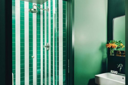 A modern shower with green and white striped tiles, a sleek showerhead, and a small sink with flowers nearby.