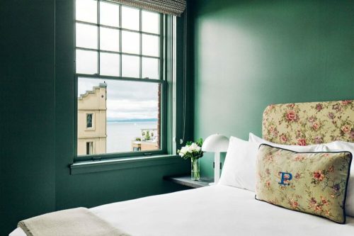 Cozy hotel room with a floral headboard, a window view of the sea, and a small vase of flowers on the bedside table.