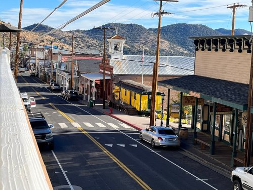 A quiet street in a small town, lined with shops and cars, with mountains in the background under a clear blue sky.