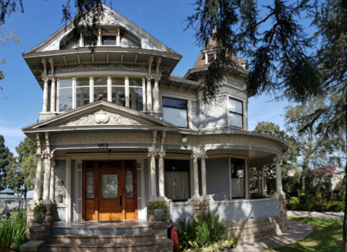 A large, ornate Victorian-style house with a wraparound porch and intricate architectural details, surrounded by greenery.