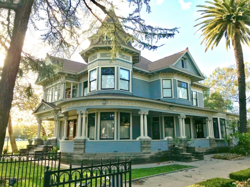 A large, ornate Victorian house with a wraparound porch, surrounded by trees and palm plants.