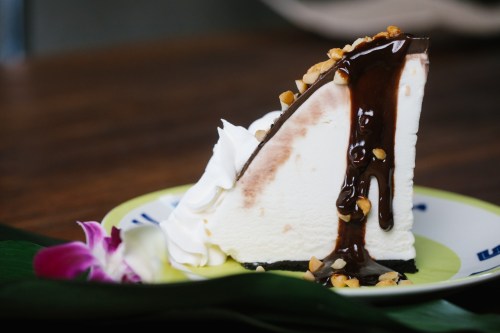 A slice of cheesecake topped with chocolate sauce, nuts, and whipped cream, served on a decorative plate with a flower.