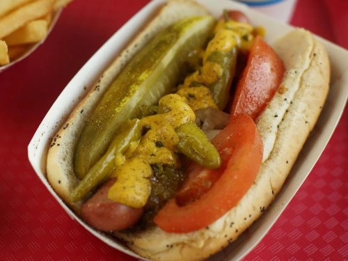 A hot dog in a bun topped with pickles, mustard, green peppers, and tomato slices, served in a paper tray.