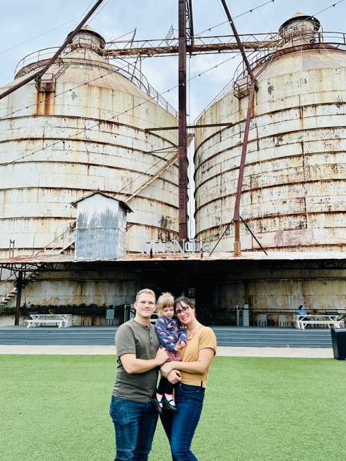 A family of three stands in front of large, rusty silos, with green grass in the foreground and a cloudy sky above.