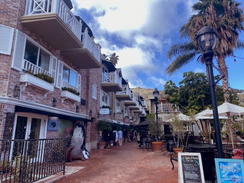 A charming brick walkway lined with shops and palm trees, featuring balconies and outdoor seating under a blue sky.