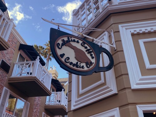 Sign for Catalina Coffee & Cookie Co. hanging from a building, with a coffee cup design and blue sky in the background.