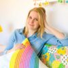 A woman with blonde hair sits on a colorful couch, holding a rainbow-patterned pillow and smiling at the camera.