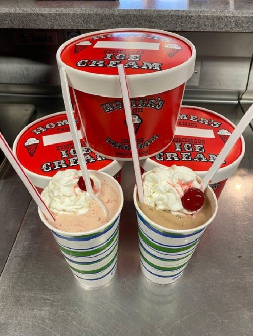 Two milkshakes topped with whipped cream and cherries, with tubs of red "Homer's Homemade Ice Cream" in the background.