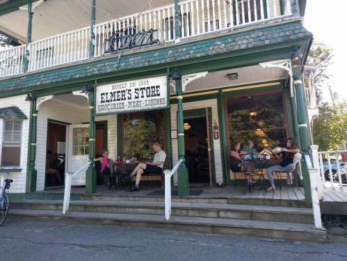 A quaint store with a sign reading "Elmer's Store," featuring outdoor seating and patrons enjoying their time.