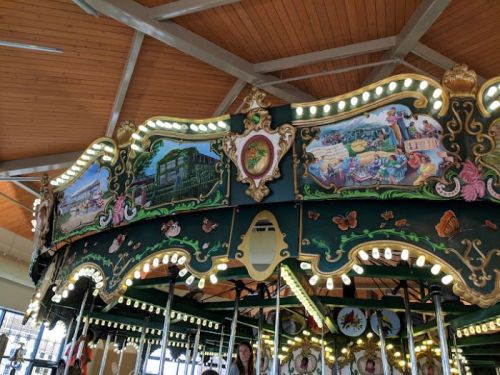 A colorful carousel with ornate decorations, featuring painted scenes and lights, under a wooden ceiling.