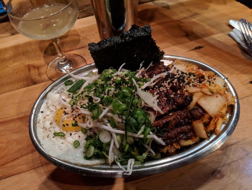 A metal bowl filled with rice, grilled meat, vegetables, a fried egg, kimchi, and a piece of seaweed, with a drink beside it.