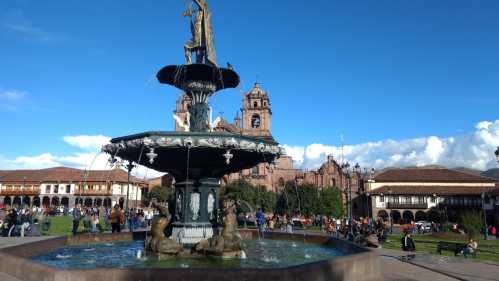 A fountain in a plaza with people, surrounded by historic buildings and a blue sky in Cusco, Peru.