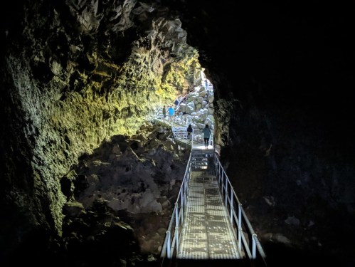A dimly lit cave with a metal walkway leading through rocky terrain, people walking in the distance.