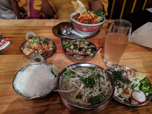 A table set with various dishes: rice, salad, noodles, and appetizers, alongside a glass of beverage.