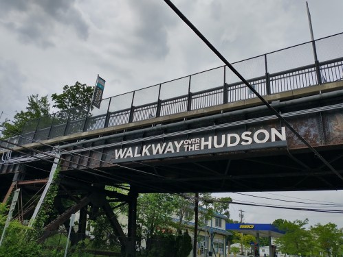A bridge with a sign reading "WALKWAY OVER THE HUDSON," surrounded by trees and a cloudy sky.