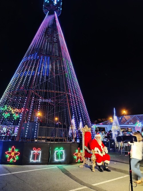 Santa Claus sits on a throne in front of a brightly lit Christmas tree at night, surrounded by festive decorations.