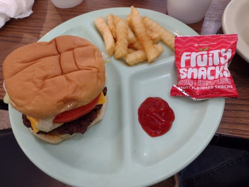 A plate with a cheeseburger, crinkle-cut fries, ketchup, and a pack of fruity snacks.
