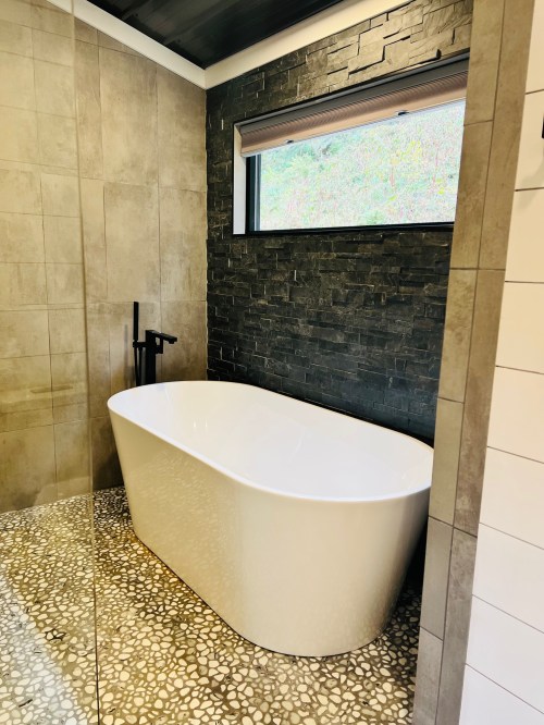 Modern bathroom featuring a freestanding white bathtub, stone wall accents, and a pebble floor. Natural light from a window.