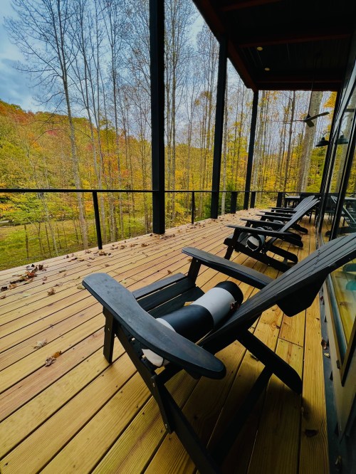 A wooden deck with black chairs overlooks a forest of autumn trees under a cloudy sky.