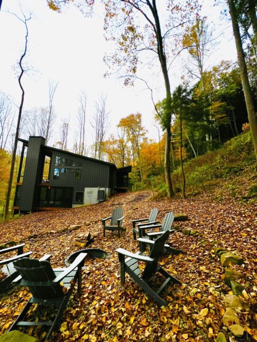 A modern cabin surrounded by autumn foliage, with Adirondack chairs arranged around a fire pit on a leaf-covered ground.