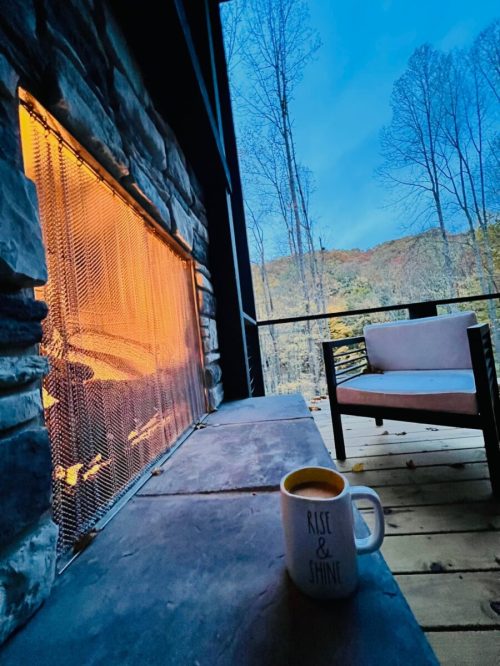 A cozy outdoor scene featuring a stone fireplace, a mug with "Rise & Shine," and a view of trees and mountains.