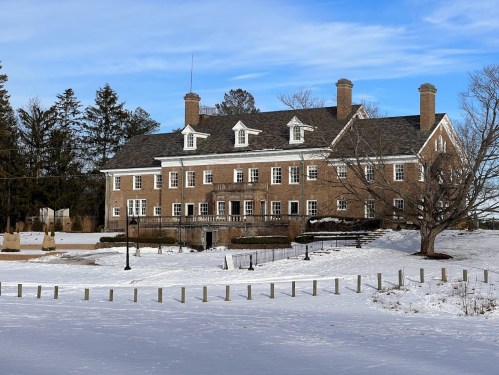 A large brick building with multiple windows, surrounded by snow and trees under a clear blue sky.