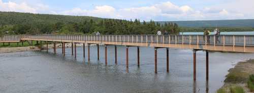 A wooden bridge spans a river, with people walking and lush greenery in the background under a partly cloudy sky.