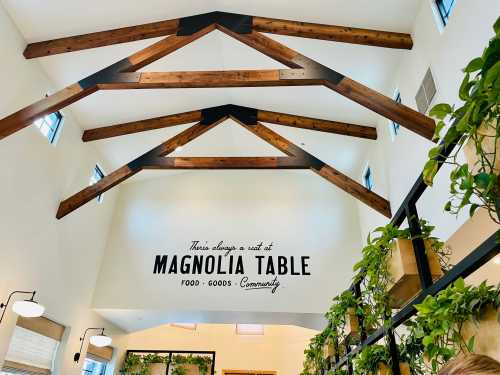 Interior of a bright space with wooden beams and a sign that reads "Magnolia Table - Food, Goods, Community."