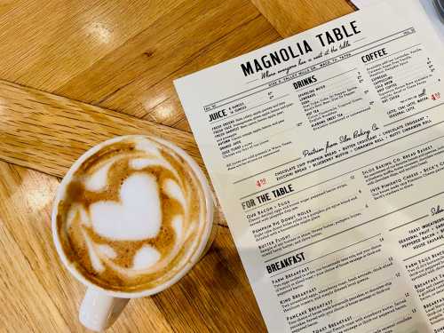 A latte with heart-shaped foam art beside a menu for Magnolia Table on a wooden table.