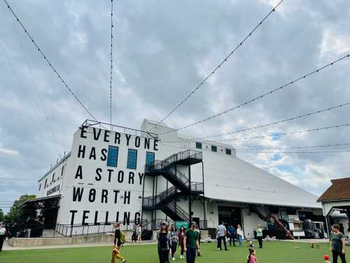 A large building with the words "Everyone has a story worth telling" on its side, surrounded by people and string lights.