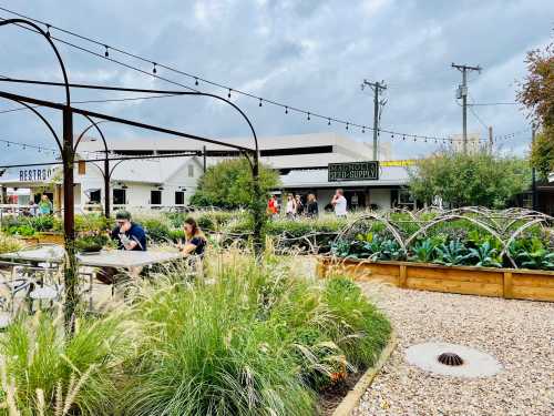 A garden area with tables, string lights, and people walking near a building labeled "Bacon & Beer Supply."